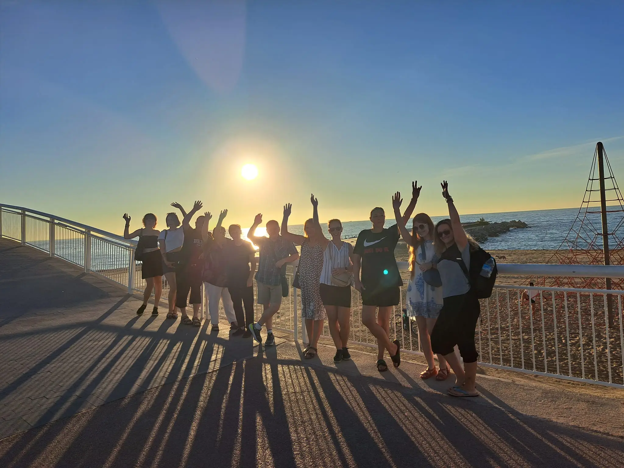 Skupina dospělých studentů španělštiny na zájezdu do Barcelonu, fotí se na pláži za východu slunce, mávají na kameru.